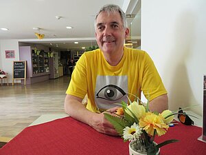 Harry R. ist sehr dankbar, dass ihm die Hilfsgemeinschaft hilft, ein selbstständiges Leben zu führen Harry sitzt mit einem gelben T-Shirt der Hilfsgemeinschaft an einem Tisch und lächelt.