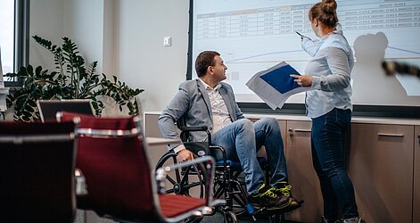 Eine Frau zeigt einem Mann im Rollstuhl eine Tabelle mit Zahlen, die auf einer Beamer-Leinwand angezeigt wird.