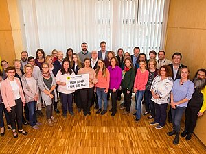 Wir sind für Sie da, Copyright: Franz Pfluegl Ungefähr 40 Menschen stehen im Halbkreis nebeneinander. in einem Raum Ganz vorne stehen zwei Frauen mit einem Schild mit der Aufschrift "Wir sind für Sie da".