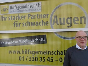 Daniele M. setzt sich als Mitarbeiter der Hilfsgemeinschaft seit 2005 für Barrierefreiheit ein. Mitarbeiter Daniele M. steht vor einem gelbschwarzen Plakat der Hilfsgemeinschaft