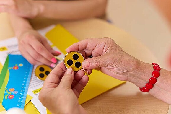 Nahaufnahme der Hände zweier Personen, die einen gelben Button mit drei schwarzen Punkten betrachten. Eine Person hält den Button. Er ist aus Metall. Auf dem Tisch liegt ein weiterer Button. 