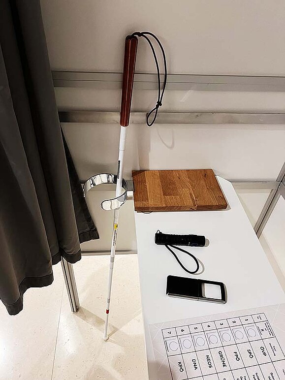 Inneres einer barrierefreien Wahlzelle: am Rand ist eine runde Halterung für Gehhilfen/Weißen Stock angebracht, in der ein Weißer Stock lehnt. Am Tisch liegen eine Taschenlampe, eine Lupe und eine Stimmzettelschablone.
