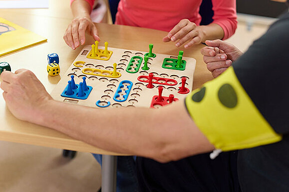 Zwei Personen spielen Mensch ärgere dich nicht. Eine Person trägt eine gelbe Armbinde mit schwarzen Punkten. Das Spielbrett hat Vertiefungen für die Figuren und die Würfelaugen sind erhaben, damit man die gewürfelte Zahl auch fühlen kann. 