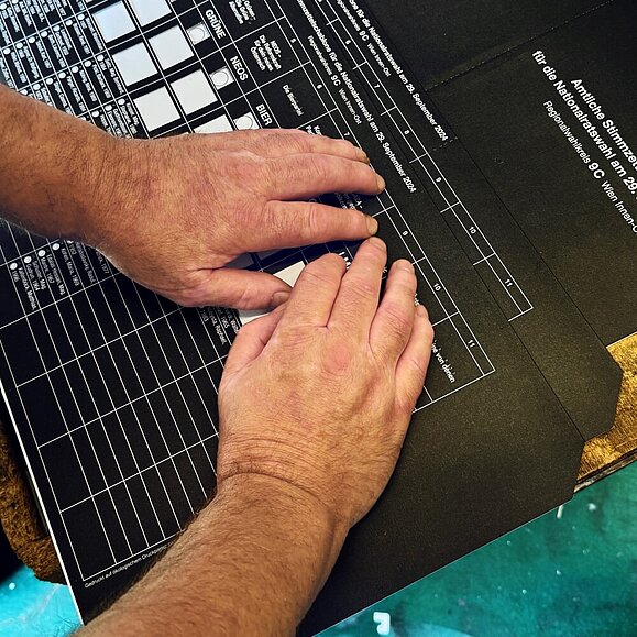 Männerhände lesen die Braille-Beschriftung auf einer schwarzen Wahlschablone, die auf einem Tisch liegt.