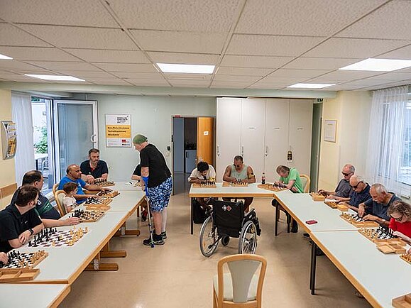 In einem Gemeinschaftsraum sitzen mehrere Personen an U-förmig gestellten Tischen und spielen simultan Schach. Eine Person mit Krücke geht von Brett zu Brett und spielt gegen alle anderen. In der Mitte steht ein leerer Rollstuhl. Die Atmosphäre ist konzentriert und ruhig.