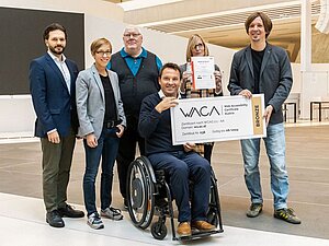 Gruppe von Menschen mit Anzug lächeln in die Kamera. Einer sitzt im Rollstuhl und hält einen überdimensionalen Scheck mit Aufschrift: WACA Zertifikat - Bronze