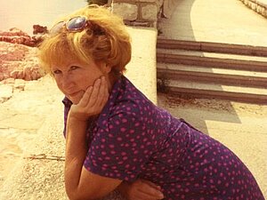 Emilia Fit Auf einem leicht vergilbten Foto sieht man eine Frau in einer mediterranen Landschaft am Meer, an einer Mauer lehnend in die Kamera lächeln.