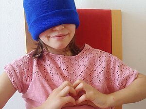 Der Blue Beanie Day, der Tag der blauen Mütze, steht für barrierefreies Internet. Ein Mädchen mit einer blauen Mütze auf dem Kopf und vor den Augen formt mit ihren Händen ein Herz
