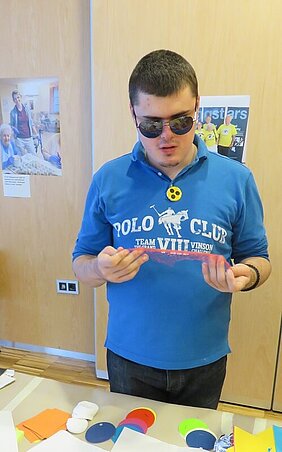 Unser Sehsternchen Dusan beim Erforschen der unterschiedlichen Materialien. Jugendlicher ertastet ein rotes Obstnetz. Er trägt eine dunkle Sonnenbrille und eine gelbe Plakte mit drei schwarzen Punkten.