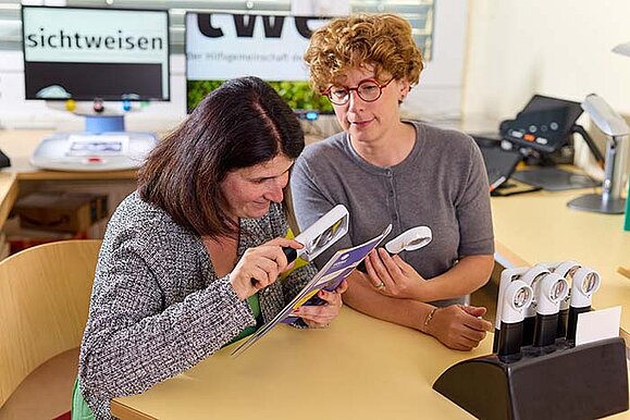 Zwei Frauen sitzen an einem Tisch in einem Büro und betrachten gemeinsam ein Heft. Die eine Frau blickt dabei durch eine Lupe, die andere beobachtet aufmerksam und hält eine weitere Lupe in der Hand. Auf dem Tisch steht ein Halter mit vielen anderen Lupen. Im Hintergrund sind Lesegeräte, auf einem ist groß die Aufschrift "Sichtweisen" zu sehen.