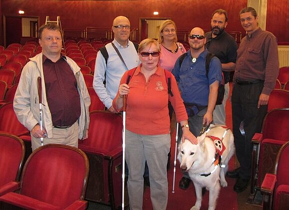 Zuschauergruppe nach einer audiokommentierten Vorstellung im Burgtheater. (c) AUDIO2 Gruppe von Menschen in rotem Theatersaal teils mit Brillen, Langstock und vorne Frau mit Blindenführhund