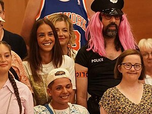 Filmausschnitt: Eine Gruppe Menschen steht da und blicken auf etwas, während sie lächeln. Manche von ihnen haben das Down-Syndrom, ein Mann mit Bart trägt eine rosarote Perücke und ein Polizeikostüm, ein anderer, sehr großer Mann im Hintergrund ein Basketballtrikot. 