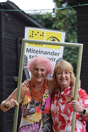 Zwei Frauen halten einen Bilderrahmen, hinter ihnen die Pappfigur vom Maskottchen AUGust