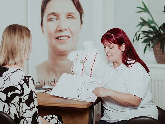 Zwei Frauen sitzen an einem Tisch, eine davon erklärt der anderen etwas anhand eines Folders.