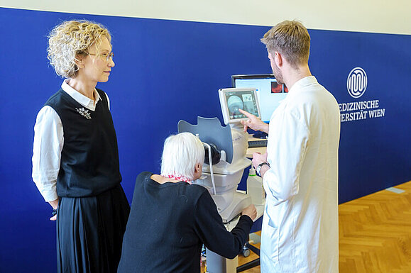 Klinikleiterin Ursula Schmidt-Erfurth und Kostiantyn Lupyr von der Universitätsklinik für Augenheilkunde und Optometrie mit einer Patientin, Copyright: MedUni Wien/Kovic Eine ältere Frau schaut in ein Gerät, dass von einem jungen Herrn bedient wird. Daneben steht eine blonde Frau.
