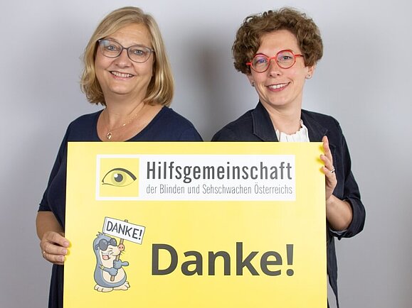 Danke, dass Sie für unsere hochgradig sehbehinderten und blinden Mitglieder da sind! Zwei Mitarbeiterinnen der Hilfsgemeinschaft lächeln und halten ein gelbes Schild hoch, auf dem groß Danke steht