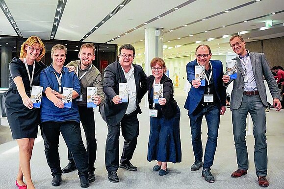 Bei der IAAP-Tagung kommen Barrierefreiheitsexperten aus der ganzen Welt zusammen. 7 Menschen in Businesskleidung und mit Umhängeschild strecken Flyer mit Aufschrift "IAAP" in die Kamera.