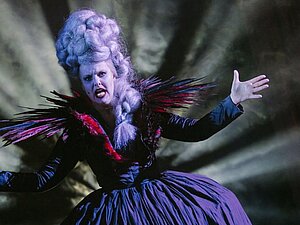 verkleidete pompöse Frau mit weißer Perücke und weiß gepudertem Gesicht, pompöses Kleid, dahinter Scheinwerfer