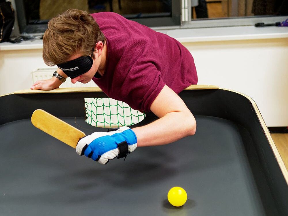 Junger Mann mit purpur T-Shirt, Handschuhen und Augenbinde spielt Tennisball auf Tisch mit länglichem Schläger