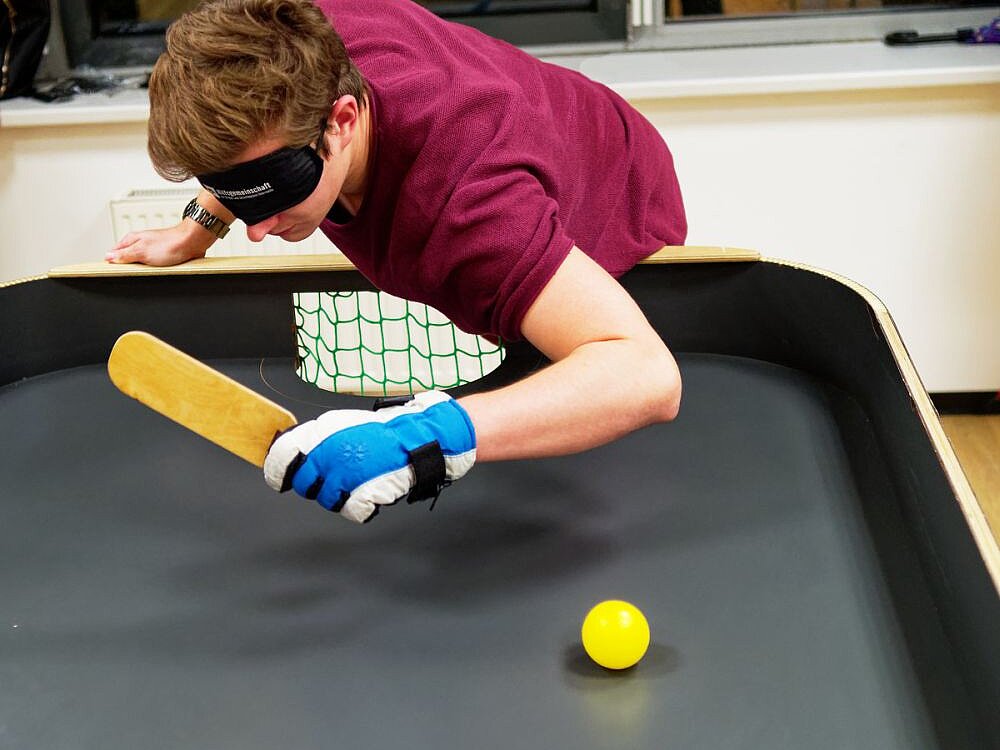 Junger Mann mit purpur T-Shirt, Handschuhen und Augenbinde spielt Tennisball auf Tisch mit länglichem Schläger