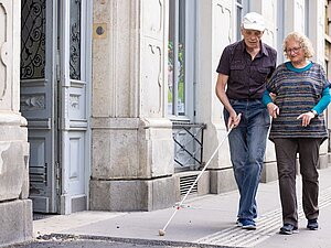 Frau mit grauen Haaren begleitet Mann mit Langstock am Gehsteig, Apothekenzeichen im Hintergrund 