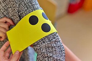 Eine Blindenschleife (drei schwarze Punkte auf gelben Hintergrund) wird auf einem Oberarm befestigt